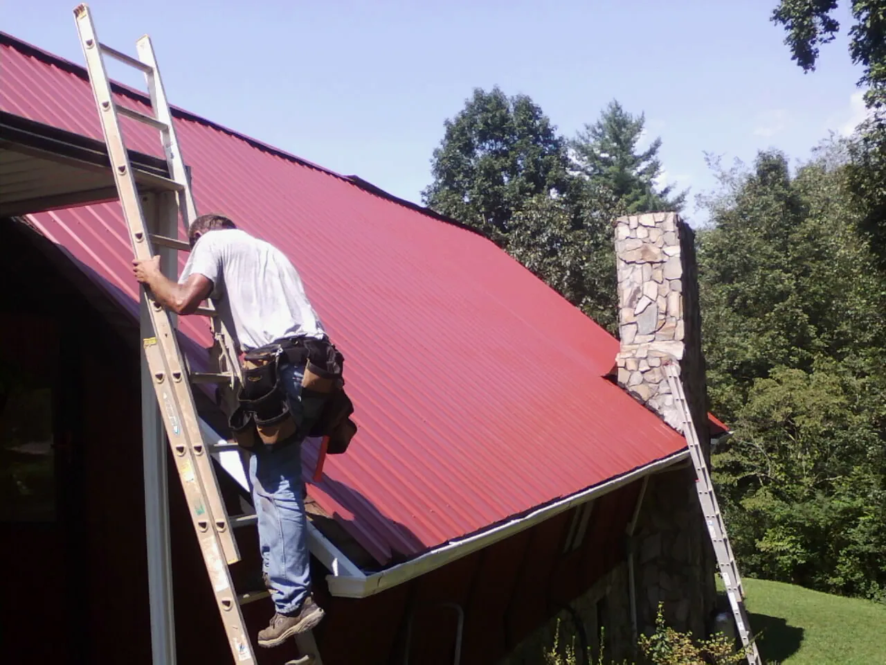 Roofer on a ladder beside a red metal roof and stone chimney — Soffit & Fascia Repair by Roofing Brothers in East Liverpool